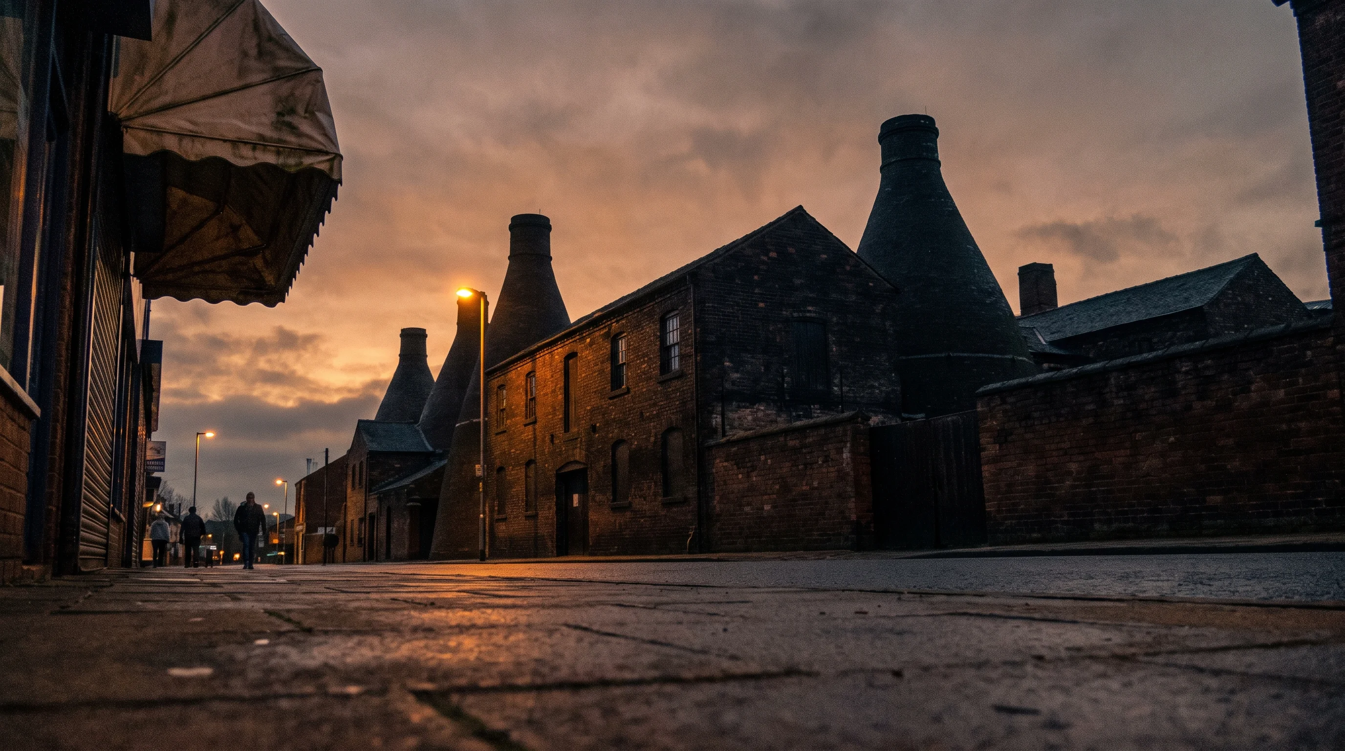 The Gladstone Pottery Museum's bottle kilns rising from soot-black brick at dusk, seen from the pavement, with the edge of a shop awning at the side of the frame